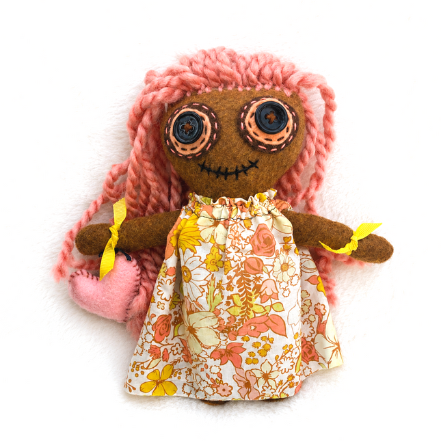 Handmade Hoodoo doll with pink hair, floral dress, and yellow bows on a white background